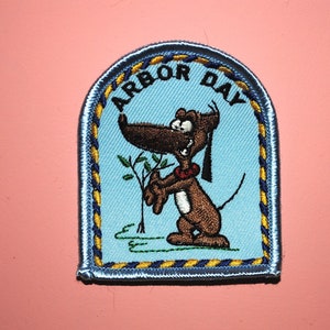 May include: A blue and yellow embroidered patch with the text "Arbor Day" and a cartoon dog holding a small tree.
