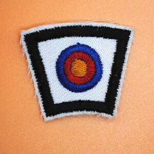 May include: A white fabric patch with a black border. The patch features a red, orange, and blue target design.
