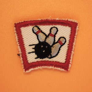 May include: A red and white embroidered patch with a bowling pin design. The patch is shaped like a square with rounded corners.