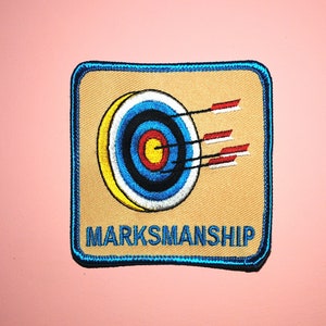 May include: A blue and tan embroidered patch with a target and arrows. The text "MARKSMANSHIP" is below the target.