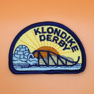 May include: A yellow and blue embroidered patch with the text "KLONDIKE DERBY" in a circular shape. The patch features a stylized image of an igloo, a bridge, and a sunrise.