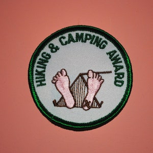 May include: A green and white embroidered patch with the text "Hiking & Camping Award" and a cartoon image of two feet sticking out of a tent.