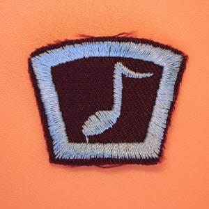 以下が含まれることがあります： 濃い赤褐色の布地に、薄い青色の縁取りと薄い青色の音符が中央に描かれたパッチ