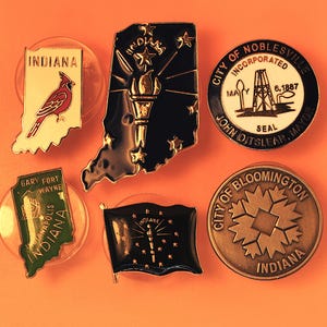 May include: Six enamel pins featuring Indiana themes. One is a cream-colored Indiana state shape with a red cardinal. One is a black Indiana shape with a gold torch and stars. One is a round Noblesville seal. One is a green Indiana shape. One is a black Indiana flag pin. One is a round bronze Bloomington seal.