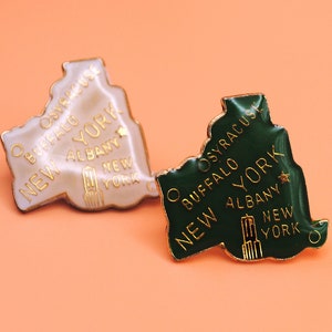 May include: Two enamel pins shaped like the state of New York. One pin is white with gold lettering and the other is green with gold lettering. Both pins say "New York" and list cities including "Buffalo, Syracuse, Albany".