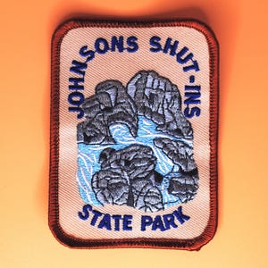 May include: Embroidered patch featuring a landscape scene with the text "JOHNSONS SHUT-INS STATE PARK". The patch has a rectangular shape with rounded corners, a tan background, and a brown border. The design includes gray rock formations and blue water.