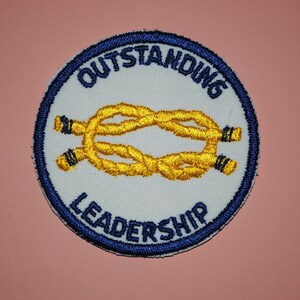 May include: A white embroidered patch with a blue border. The patch features a yellow rope knot and the text "OUTSTANDING LEADERSHIP".