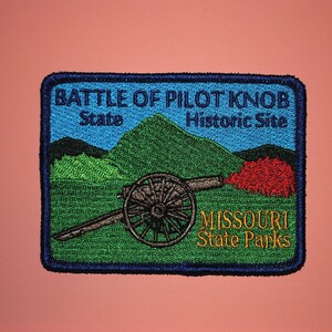May include: A blue and green embroidered patch with the text "Battle of Pilot Knob State Historic Site" and "Missouri State Parks". The patch features a cannon with a red plume of smoke.