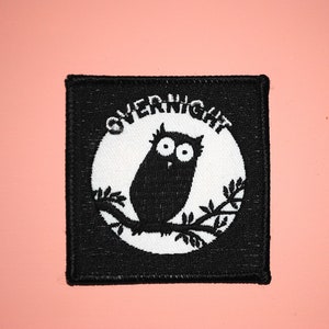 May include: Black and white embroidered patch with an owl perched on a branch inside a circle. The circle has the word "OVERNIGHT" written around it.