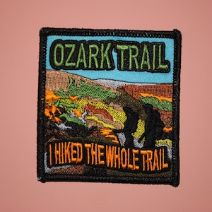 May include: A black and green embroidered patch with a blue background. The patch features a landscape scene with two hikers and the text "Ozark Trail" and "I hiked the whole trail".