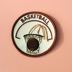 May include: A brown and white embroidered patch with the word "BASKETBALL" in brown lettering around the edge. The center of the patch shows a brown basketball going through a brown hoop with a white net.