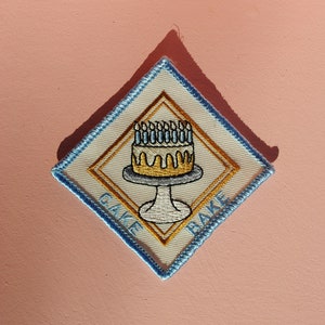May include: A white diamond-shaped patch with a blue border. The patch features a yellow cake with blue candles and the words "Cake Bake" in gold embroidery.
