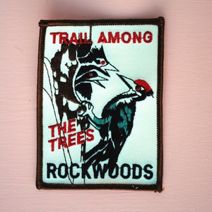 May include: A brown embroidered patch with white background featuring a black and green bird with red accents. The text "TRAIL AMONG THE TREES ROCKWOODS" is embroidered in red.