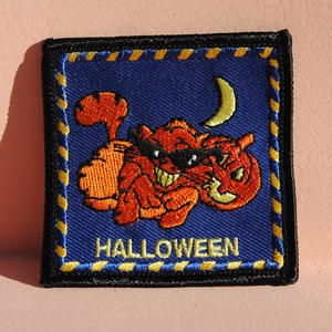 May include: Square patch with a Halloween theme. The patch features an orange monster wearing sunglasses and holding a jack-o'-lantern. A yellow crescent moon is in the background. The word "HALLOWEEN" is embroidered in yellow.