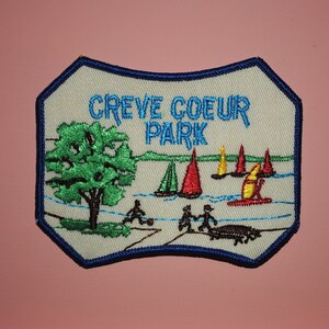 以下が含まれることがあります： 青い縁取りのある白い刺繍パッチで、"Creve Coeur Park"という文字と、緑の木と岸辺の人々がいる湖に帆船が浮かぶ風景が描かれています。