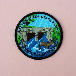 以下が含まれることがあります： 青い水と緑の草木が刺繍された、石の橋が架かっている丸い黒い縁取りのパッチ。橋の下には鹿が水の中にいる。パッチの周りに白い円形で「Knob Noster State Park」という文字が書かれており、その下に白い弧状に「Knob Noster, Missouri」という文字が書かれている。