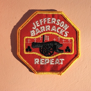 May include: Embroidered patch with a red octagonal shape and yellow trim. The patch features the text "JEFFERSON BARRACKS REPEAT" in white, along with a black cannon illustration. A flag is also visible.
