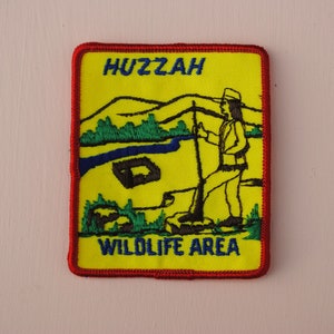 May include: A yellow embroidered patch with red stitching. The patch features a cartoon image of a person with a rifle standing in a field with a river and mountains in the background. The text "HUZZAH WILDLIFE AREA" is embroidered in blue on the patch.