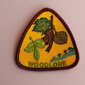 May include: A triangular yellow patch with a brown border. The patch has an embroidered design of leaves, a nut, and mushrooms. The text "WOODLORE" is embroidered in brown at the bottom of the patch.