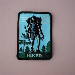 May include: A green embroidered patch with a hiker silhouette carrying a backpack and walking stick. The patch has a blue background with white clouds and green trees. The text "HIKER" is written in white at the bottom of the patch.