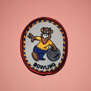May include: A red, yellow, and blue embroidered patch with a cartoon bear bowling. The patch says "BOWLING" in a white arc.