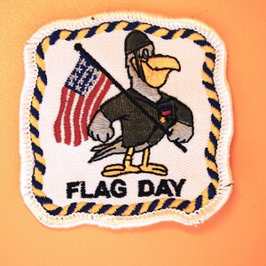 May include: Embroidered patch featuring a cartoon bird in a military uniform holding an American flag. The patch has a white background with a blue and yellow rope border and the words "FLAG DAY".