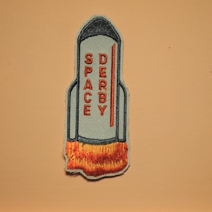 May include: A gray and orange embroidered patch shaped like a rocket with the words "SPACE DERBY" on the side.