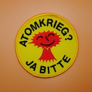 以下が含まれることがあります： 「ATOMKRIEG? JA BITTE」というテキストと、笑顔の赤いキノコ雲が描かれた黄色と黒の刺繍パッチです。