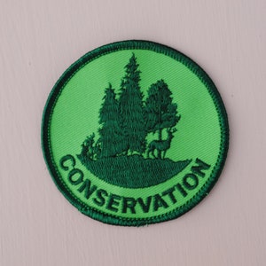 May include: Green embroidered patch with a silhouette of trees and a deer. The patch is circular and has the word "CONSERVATION" written around the edge.