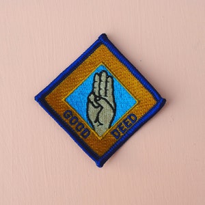 May include: A diamond-shaped patch with a blue border and gold interior. The center features a hand symbol and the words "GOOD DEED" in gold. The patch is likely for a youth organization.