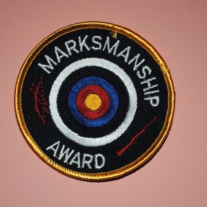 May include: A black and gold embroidered patch with a target and rifle. The text reads "Marksmanship Award".
