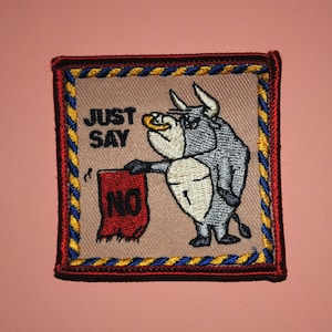 May include: A brown and red embroidered patch with a cartoon bull holding a red flag that says "NO". The bull is wearing a white shirt and has a blue and yellow border around it. The text "JUST SAY" is above the bull.