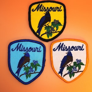 以下が含まれることがあります： 青い鳥が枝にとまっている刺繍のアイロン接着パッチ3枚。パッチには青い文字で「ミズーリ」と書かれています。パッチの色は黄色、青、白です。