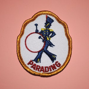 May include: A white embroidered patch with a gold border. The patch features a blue and red figure of a drummer marching with a drum. The text "PARADING" is written in red below the figure.