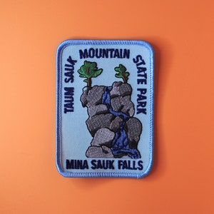 May include: A blue embroidered patch with the text "Mina Sauk Falls,  Taum Sauk Mountain State Park". The patch features a cartoon illustration of a waterfall cascading over rocks with two green trees.