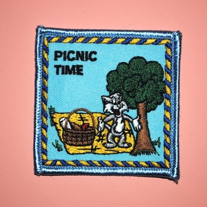 May include: A blue and yellow embroidered patch with a cartoon cat holding a picnic basket. The text "PICNIC TIME" is written in black letters on the patch.