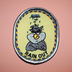 以下が含まれることがあります： 黒い帽子とサングラスをかけた漫画の鳥が描かれた黄色と青の刺繍パッチ。鳥は眉をひそめて下を向いており、鳥の下には「RAIN OUT」という文字が書かれている。
