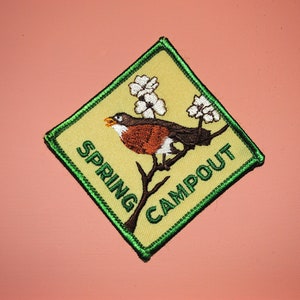 May include: A yellow diamond-shaped patch with a green border. The patch features a robin perched on a branch with white blossoms. The text "SPRING CAMPOUT" is embroidered in green on the patch.
