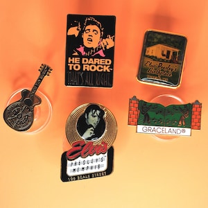 May include: A collection of Elvis Presley themed pins and magnets. Includes a guitar, a black and white image of Elvis, and pins with text that reads "He Dared to Rock", "Elvis Presley's Birthplace", and "Graceland".