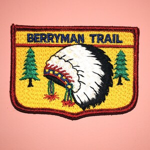 May include: A yellow and red embroidered patch with the text "BERRYMAN TRAIL" in blue. The patch features a stylized Native American headdress with white feathers and a black and red band. Two green pine trees are on either side of the headdress.
