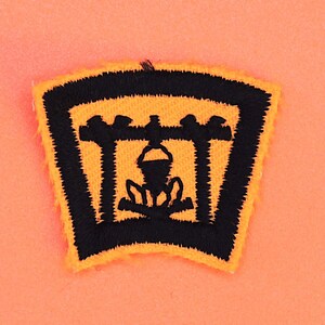 May include: A black and yellow embroidered patch with a campfire scene. The patch features a campfire with a pot hanging over the flames, surrounded by a black frame.