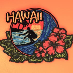 May include: Embroidered patch featuring a surfer riding a wave with a sunset backdrop. The word "HAWAII" is in orange letters above the scene. Red hibiscus flowers and green leaves frame the design, set against an orange background.