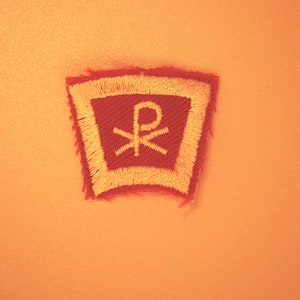 May include: A red and yellow embroidered patch with a white symbol, the Chi Rho, a Christian symbol.