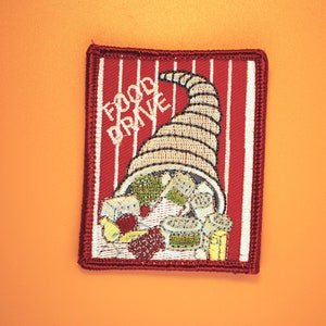 以下が含まれることがあります： 赤い白の布のパッチで、食べ物が溢れる豊穣の角と「フードドライブ」の文字が書かれています。