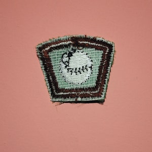 Pu&ograve; includere: Una toppa ricamata verde e marrone con una palla da baseball bianca al suo interno. La palla da baseball ha cuciture e un piccolo punto nero al centro.