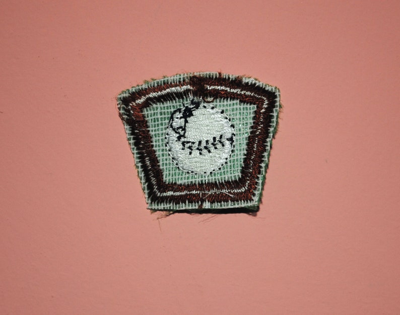 Pu&ograve; includere: Una toppa ricamata verde e marrone con una palla da baseball bianca al suo interno. La palla da baseball ha cuciture e un piccolo punto nero al centro.