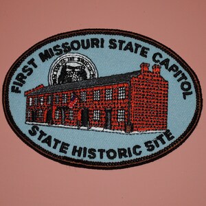 May include: A blue and black oval patch with the text "First Missouri State Capitol State Historic Site" and a red brick building with a white flag.
