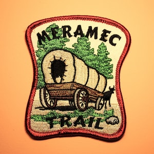 May include: A red, white, and green embroidered patch with the text "Meramec Trail" and a depiction of a covered wagon pulled by an ox.