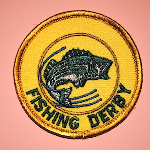 May include: A yellow and gold embroidered patch with a green and white fish jumping out of the water. The text "Fishing Derby" is written around the fish.
