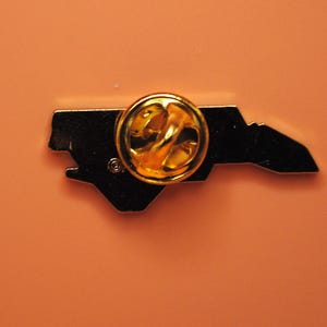 May include: A black enamel pin in the shape of the state of North Carolina, with a gold-coloured pin back. The pin is set against an orange background.